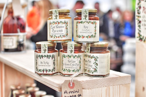 Several jars of curry paste and chutney are displayed on a wooden shelf. The labels feature decorative borders with text such as 'East Yorkshire Piccalilli', 'Ruby Curry Paste', and 'Mango, Methi & Fruit Chutney'. The jars are neatly arranged, and more bottles can be seen in the background amidst a blurred crowd.