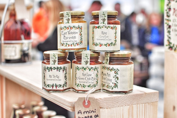 Several jars of curry paste and chutney are displayed on a wooden shelf. The labels feature decorative borders with text such as 'East Yorkshire Piccalilli', 'Ruby Curry Paste', and 'Mango, Methi & Fruit Chutney'. The jars are neatly arranged, and more bottles can be seen in the background amidst a blurred crowd.