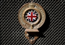 An emblem with the Union Jack in the center, flanked by wings, and inscribed with 'Royal Automobile Club Associate'. The emblem is mounted on a dark mesh background.