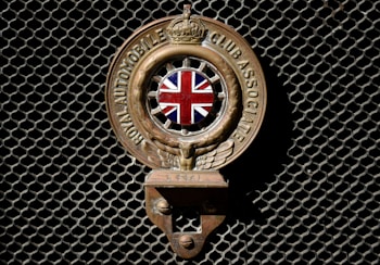 An emblem with the Union Jack in the center, flanked by wings, and inscribed with 'Royal Automobile Club Associate'. The emblem is mounted on a dark mesh background.