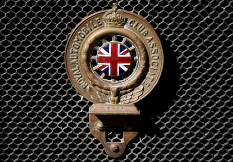 An emblem with the Union Jack in the center, flanked by wings, and inscribed with 'Royal Automobile Club Associate'. The emblem is mounted on a dark mesh background.