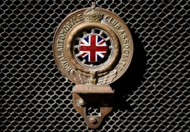An emblem with the Union Jack in the center, flanked by wings, and inscribed with 'Royal Automobile Club Associate'. The emblem is mounted on a dark mesh background.