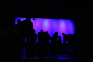 Silhouettes of people are set against a bright blue background, possibly suggesting a stage or performance setting. The figures are in various poses, with some standing and others appearing to kneel or bend.