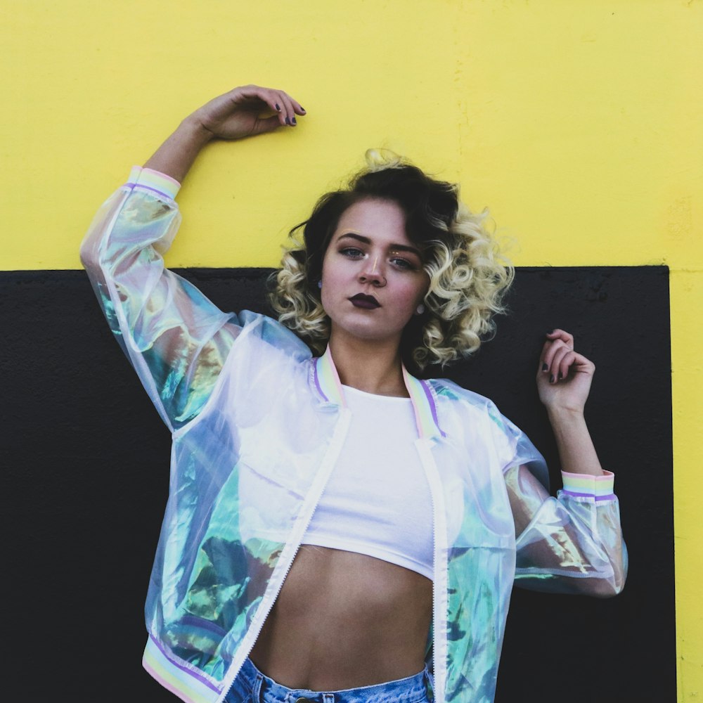 woman wearing clear plastic bomber jacket, white crop shirt, and blue denim bottoms standing behind yellow and black painted wall