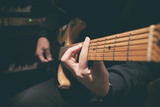 A person playing an electric guitar, with one hand positioning chords on the guitar neck. The focus is on the strings and the fretboard, while the background shows an amplifier, suggesting a musical setting.
