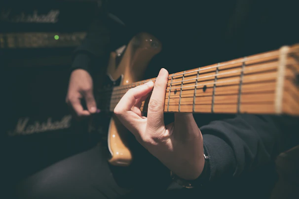 A person playing an electric guitar, with one hand positioning chords on the guitar neck. The focus is on the strings and the fretboard, while the background shows an amplifier, suggesting a musical setting.