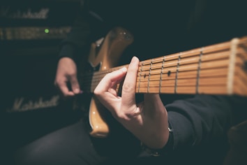 A person playing an electric guitar, with one hand positioning chords on the guitar neck. The focus is on the strings and the fretboard, while the background shows an amplifier, suggesting a musical setting.