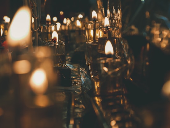 A multitude of small candle flames illuminating the surroundings with a warm, golden glow. The reflections on glass surfaces create a soft, flickering ambiance, suggesting a serene and contemplative atmosphere. The blurred background enhances the foreground focus on the delicate candlelight.
