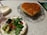 A heart-shaped cake with a caramelized top is presented on a decorative glass plate. Next to it is a plate with a fresh green salad, including cherry tomatoes, cucumber, and sliced avocado, on a vintage-style floral plate. The setting is on a lace tablecloth, creating a cozy dining atmosphere.