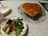 A heart-shaped cake with a caramelized top is presented on a decorative glass plate. Next to it is a plate with a fresh green salad, including cherry tomatoes, cucumber, and sliced avocado, on a vintage-style floral plate. The setting is on a lace tablecloth, creating a cozy dining atmosphere.