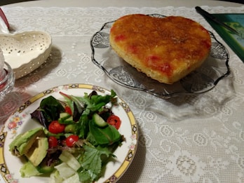 A heart-shaped cake with a caramelized top is presented on a decorative glass plate. Next to it is a plate with a fresh green salad, including cherry tomatoes, cucumber, and sliced avocado, on a vintage-style floral plate. The setting is on a lace tablecloth, creating a cozy dining atmosphere.