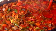 A colorful dish of cooked vegetables in a rich red sauce, featuring visible pieces of bell peppers, eggplant, and tomatoes. A wooden ladle is partially submerged in the mixture, adding to the vibrant and wholesome aesthetic of the meal.