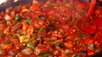 A colorful dish of cooked vegetables in a rich red sauce, featuring visible pieces of bell peppers, eggplant, and tomatoes. A wooden ladle is partially submerged in the mixture, adding to the vibrant and wholesome aesthetic of the meal.