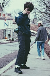 Two young individuals are on a sidewalk, one making a peace sign with their hand and wearing a dark jacket with sunglasses, while the other stands in the background holding a drink can. There are bare trees and an older building with flags in the background, likely during early spring or late winter.