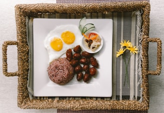 A rectangular woven tray contains a square white plate with a serving of purple rice, sunny-side-up eggs, small sausages, and a small condiment bowl with sliced vegetables. Two handles are attached to the sides of the tray, which is placed on a striped fabric mat.