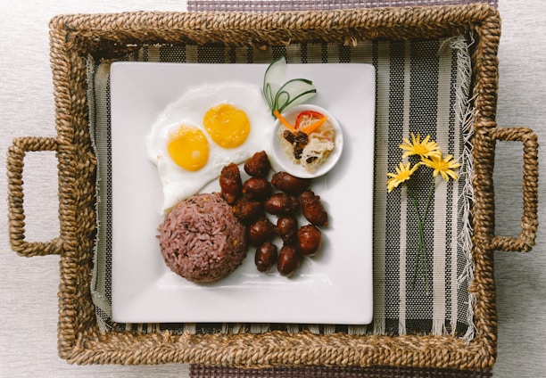 A rectangular woven tray contains a square white plate with a serving of purple rice, sunny-side-up eggs, small sausages, and a small condiment bowl with sliced vegetables. Two handles are attached to the sides of the tray, which is placed on a striped fabric mat.