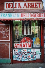 The storefront of a deli market displays various signs and advertisements. There is a Coca-Cola sign, an ATM sign, and a poster for Corona Light. The windows have additional posters, including a depiction of a woman holding a beverage. Graffiti and stickers are also visible on the door and below the windows.