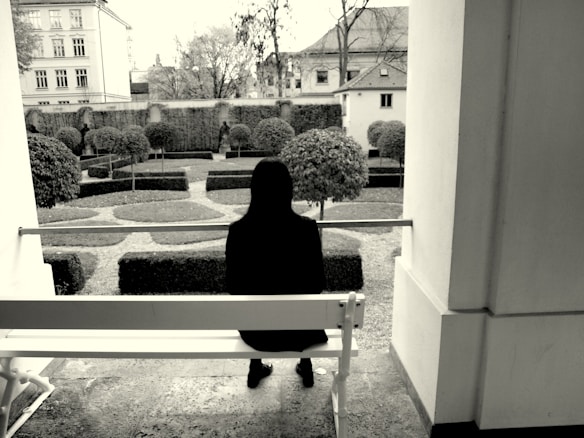 A person with long hair sits alone on a white bench in a courtyard facing a manicured garden. The scene is framed by two walls, creating a sense of solitude and introspection. The garden features neatly trimmed bushes arranged in geometric patterns, and in the background, there are several buildings and bare trees.