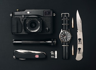 A collection of everyday carry items is arranged neatly on a black surface. The items include a black camera with a lens, a classic watch with a NATO strap, a tactical folding knife, a Swiss Army knife, an ammunition bullet, a small flashlight, and a 64GB camera memory card.