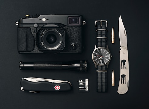 A collection of everyday carry items is arranged neatly on a black surface. The items include a black camera with a lens, a classic watch with a NATO strap, a tactical folding knife, a Swiss Army knife, an ammunition bullet, a small flashlight, and a 64GB camera memory card.