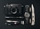 A collection of everyday carry items is arranged neatly on a black surface. The items include a black camera with a lens, a classic watch with a NATO strap, a tactical folding knife, a Swiss Army knife, an ammunition bullet, a small flashlight, and a 64GB camera memory card.