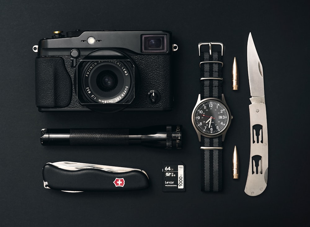 câmera preta, relógio analógico prateado redondo, canivete suíço preto e lanterna preta