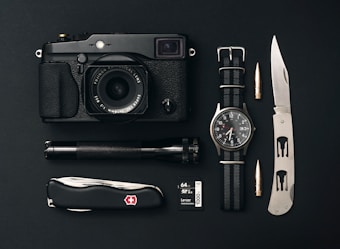 A collection of everyday carry items is arranged neatly on a black surface. The items include a black camera with a lens, a classic watch with a NATO strap, a tactical folding knife, a Swiss Army knife, an ammunition bullet, a small flashlight, and a 64GB camera memory card.