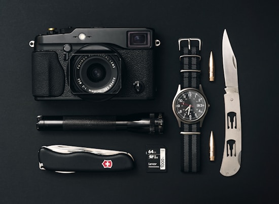 A collection of everyday carry items is arranged neatly on a black surface. The items include a black camera with a lens, a classic watch with a NATO strap, a tactical folding knife, a Swiss Army knife, an ammunition bullet, a small flashlight, and a 64GB camera memory card.