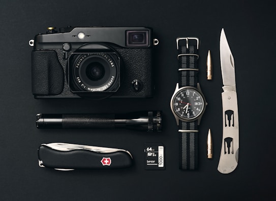 A collection of everyday carry items is arranged neatly on a black surface. The items include a black camera with a lens, a classic watch with a NATO strap, a tactical folding knife, a Swiss Army knife, an ammunition bullet, a small flashlight, and a 64GB camera memory card.