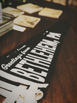 A black pennant with white lettering displays the message 'Greetings from Bethlehem, PA' on a wooden table. In the background, several square coasters or tiles and other items are slightly out of focus.