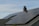 A house roof is covered with a large array of solar panels. The panels are angled to capture sunlight effectively and are bordered by a typical shingle roof. A red brick chimney protrudes through the center, indicating the presence of a fireplace or heating system inside.