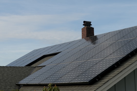 A house roof is covered with a large array of solar panels. The panels are angled to capture sunlight effectively and are bordered by a typical shingle roof. A red brick chimney protrudes through the center, indicating the presence of a fireplace or heating system inside.