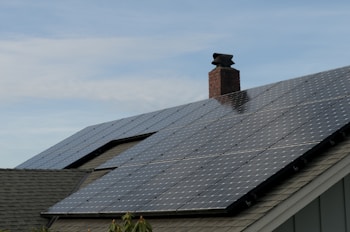 A house roof is covered with a large array of solar panels. The panels are angled to capture sunlight effectively and are bordered by a typical shingle roof. A red brick chimney protrudes through the center, indicating the presence of a fireplace or heating system inside.