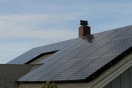 A house roof is covered with a large array of solar panels. The panels are angled to capture sunlight effectively and are bordered by a typical shingle roof. A red brick chimney protrudes through the center, indicating the presence of a fireplace or heating system inside.
