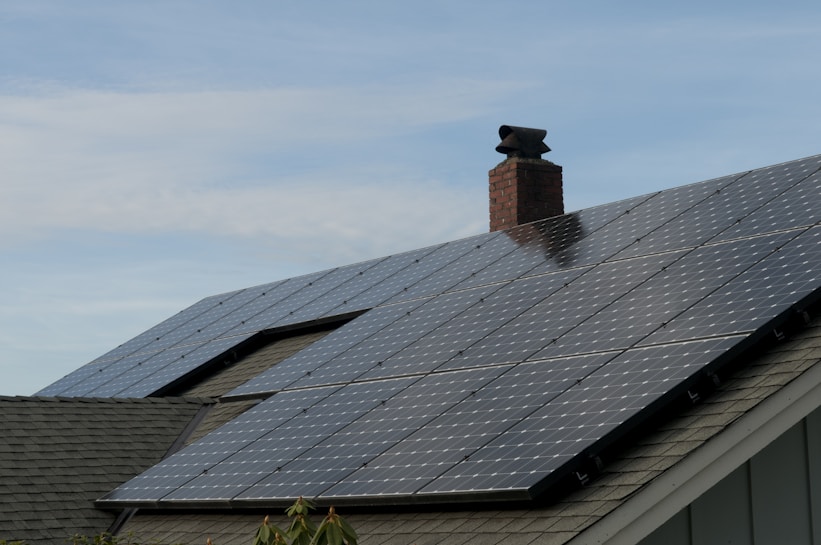 A house roof is covered with a large array of solar panels. The panels are angled to capture sunlight effectively and are bordered by a typical shingle roof. A red brick chimney protrudes through the center, indicating the presence of a fireplace or heating system inside.