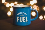 A blue enamel mug with the words 'Adventure Fuel' printed on it is placed on a dark surface, with warm, blurred lights in the background creating a cozy atmosphere.