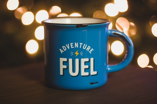 A blue enamel mug with the words 'Adventure Fuel' printed on it is placed on a dark surface, with warm, blurred lights in the background creating a cozy atmosphere.