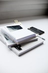 white spiral notebook beside black smartphone on white table