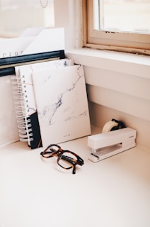 A tidy workspace featuring a stack of spiral notebooks, a marble-designed notepad, glasses, a white stapler, and a tape dispenser on a white desk. Natural light streams in through the window, illuminating the organized setup.