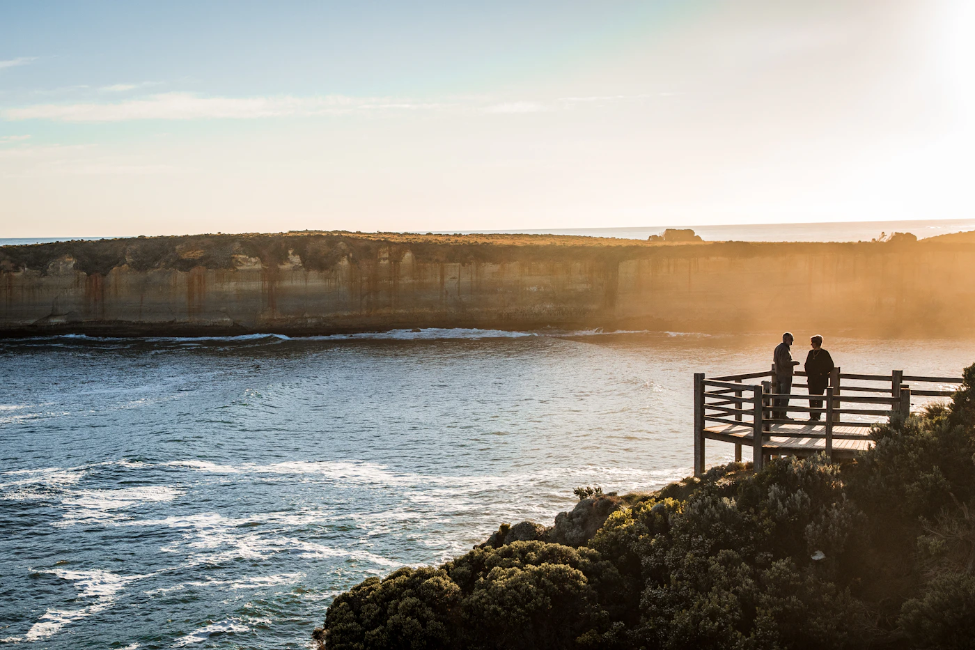 Two silhouetted figures together on a terrace overlooking vast water at golden hour with warm light on the horizon
