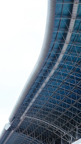 A modern architectural structure with a sweeping, curved roof made of blue and metallic materials. The intricate grid of metal beams supports transparent panels, creating a sleek, futuristic design.