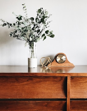 A wooden dresser with a natural finish is topped with a vase holding eucalyptus branches. Beside the vase, a decorative golden armillary sphere sculpture is placed. A wooden mantel clock with a classic design sits next to these items.