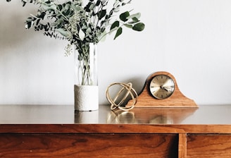 brown wooden framed analog mantle clock near gray petaled flower centerpiece