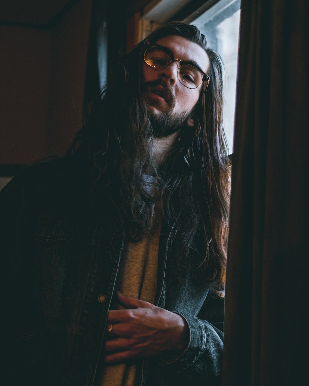 homme aux cheveux longs portant des lunettes et une veste en jean bleue appuyée sur la fenêtre