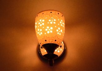 A decorative wall lamp emits a warm, orange glow. The lamp features intricate floral cut-out patterns that cast light onto a smooth, neutral-colored wall.