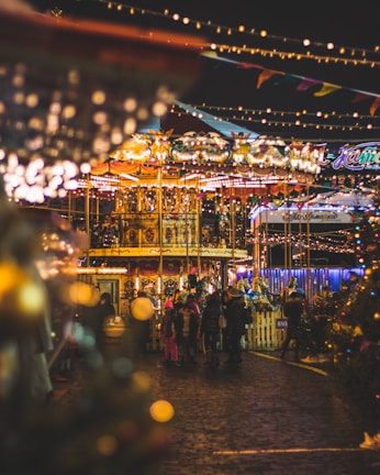 A vibrant and festive carnival scene featuring a brightly lit carousel with colorful decorations. People, including children in winter clothing, gather around the ride, creating a lively atmosphere. Warm, glowing lights and festive decorations are strung overhead, enhancing the joyful ambiance. The setting indicates an evening celebration with blurred light effects adding to the magical feel.