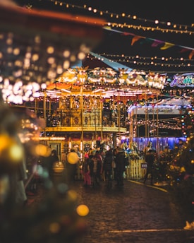 A vibrant and festive carnival scene featuring a brightly lit carousel with colorful decorations. People, including children in winter clothing, gather around the ride, creating a lively atmosphere. Warm, glowing lights and festive decorations are strung overhead, enhancing the joyful ambiance. The setting indicates an evening celebration with blurred light effects adding to the magical feel.