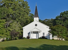 A small white church with a pointed steeple sits on a well-manicured lawn. Surrounding the building are lush green trees under a clear blue sky, creating a peaceful and serene atmosphere.