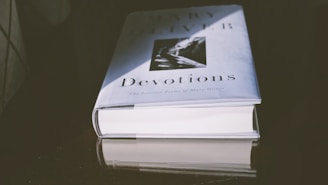 A hardcover book titled 'Devotions' with an image on the cover lays flat on a reflective dark surface. The lighting creates a shadow and highlights the title and author.