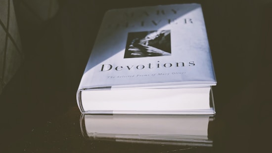A hardcover book titled 'Devotions' with an image on the cover lays flat on a reflective dark surface. The lighting creates a shadow and highlights the title and author.
